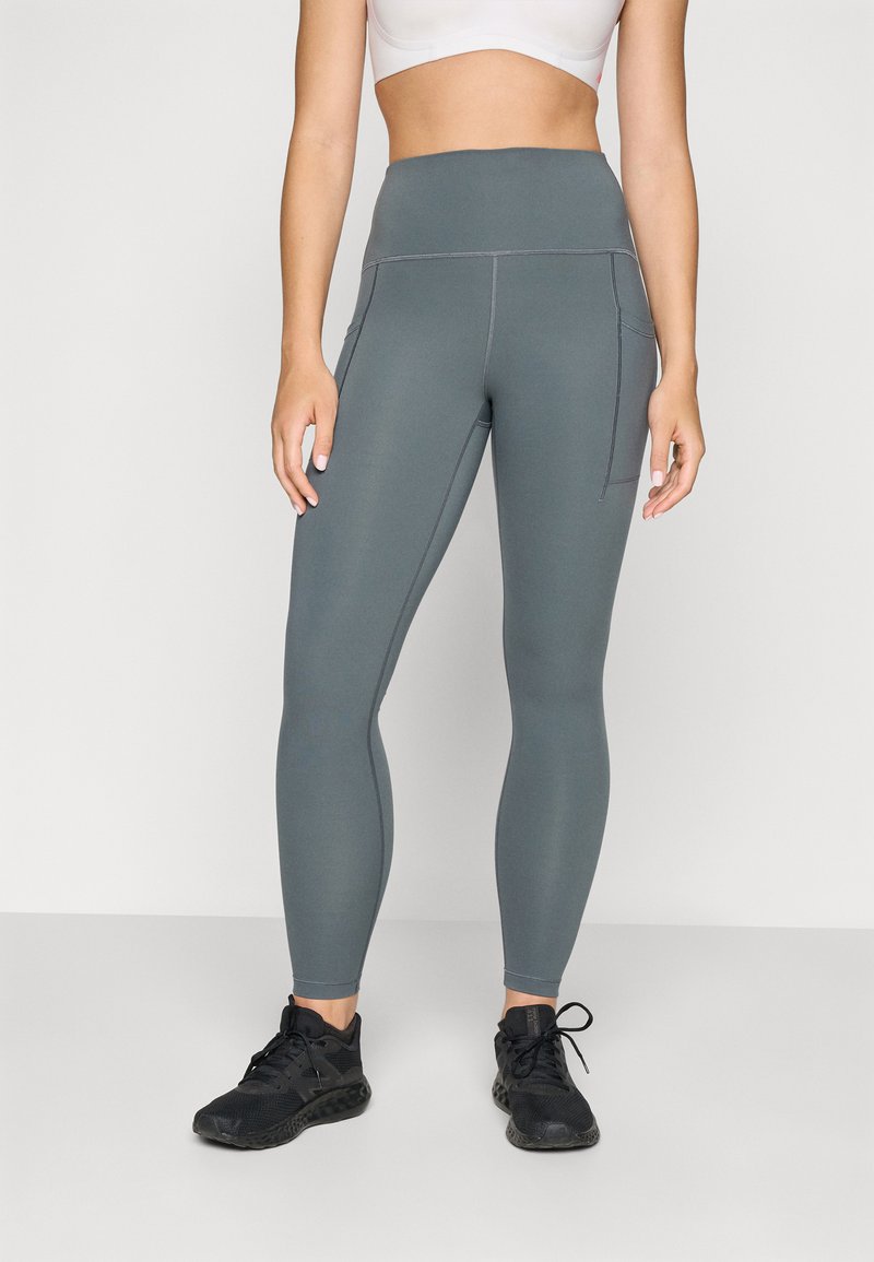 Leggings de yoga taille haute, gris avec des coutures flatlock et des poches latérales, associés à des chaussures de sport noires. Tissu extensible, design élégant.