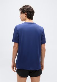 Νέος άνδρας με κοντά καστανά μαλλιά, φοράει απλό μπλε κοντομάνικο πουκάμισο και μαύρο σορτς, στραμμένος με την πλάτη προς το λευκό φόντο.