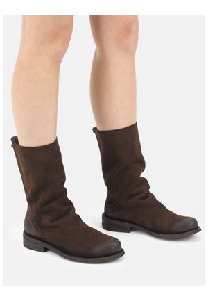 Persoon die donkerbruine suède laarzen tot halverwege de kuit draagt, met afgeronde neuzen en lage houten hakken, staand op een witte ondergrond.