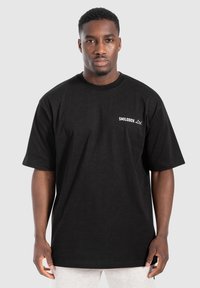 Hombre que lleva una camiseta negra de gran tamaño con el texto "SMILODOX Ltd." en el pecho, de pie contra un fondo gris claro liso.