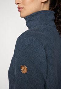 Navy fleece jacket with a high collar, featuring a tan fox logo patch on the left sleeve and smooth texture with visible stitching.