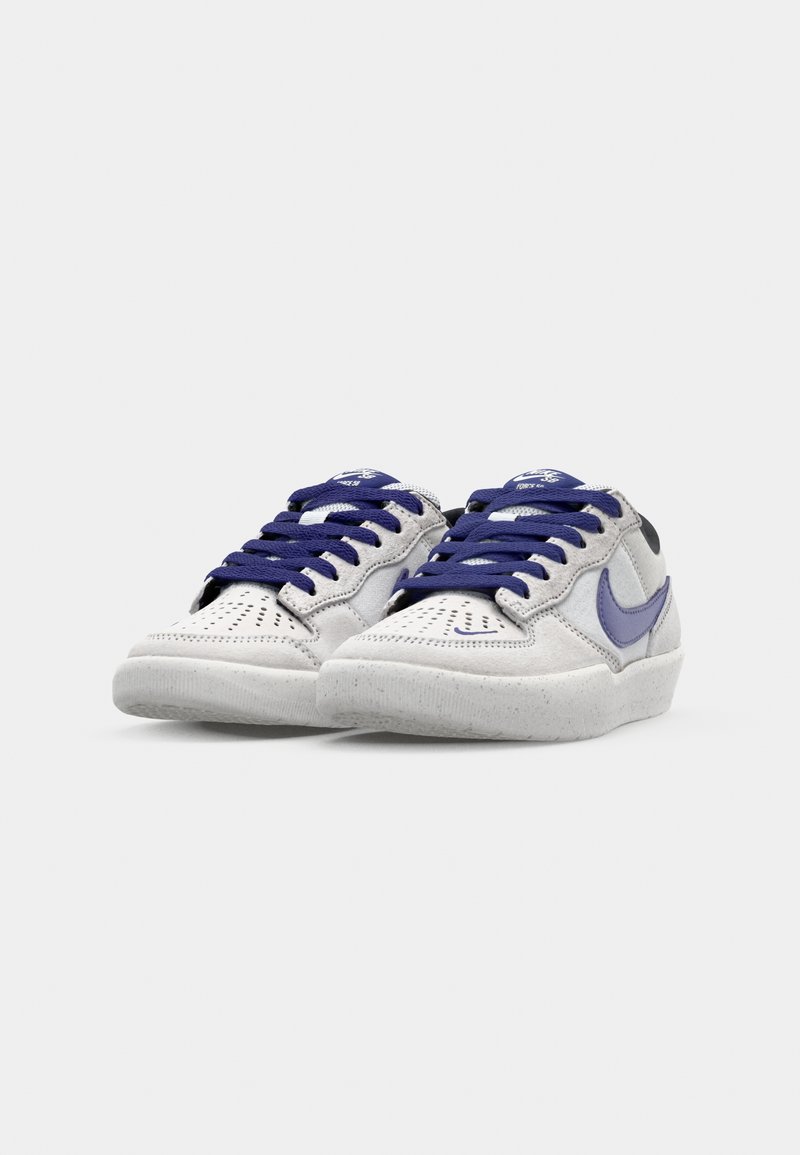 Graue Wildledersneaker mit dunkelblauen Schnürsenkeln, perforierter Zehenpartie und einem lavendelfarbenen Swoosh. Gummisohle mit subtiler Textur und leicht erhöhtem Absatz.