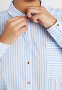 Camisa de rayas azul claro y blanca con cuello abotonado, que cuenta con botones redondos y un bolsillo en el pecho. La tela parece suave y ligera.