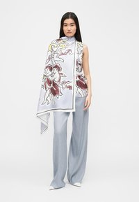 Γυναίκα που φοράει μπλούζα χωρίς μανίκια και φαρδιές ανοιχτόμπλε παντελόνες, τυλιγμένη με μεγάλο λευκό φουλάρι που έχει σχέδια με αγγελικές μορφές και φιγούρες με φτερά.