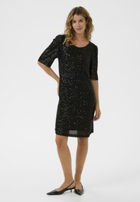 Vestido negro de lentejuelas con mangas cortas, escote redondo, corte recto y de longitud media. Llevado con tacones negros tipo slingback. Textura suave.