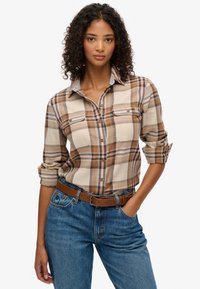 Camisa de cuadros marrón y crema con botones, dos bolsillos en el pecho, mangas remangadas y cuello gris, combinada con vaqueros azules y un cinturón marrón.