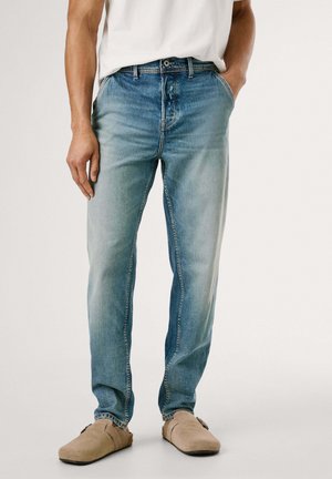 Jeans in denim blu chiaro con vestibilità dritta, caratterizzati da un lavaggio sbiadito, cinque tasche e una vita con bottone. Indossati con scarpe slip-on beige.