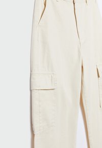 Pantalon cargo beige avec poches à rabat latérales et poches fendues à l'avant, montrant la section supérieure et médiane de la jambe sur un fond blanc.