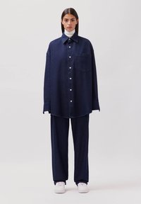 Übergroßes navy-blaues Denim-Hemd mit spitzem Kragen, Knopfverschluss und Brusttasche auf der linken Seite, kombiniert mit passenden weit geschnittenen Hosen und weißen Sneakers.