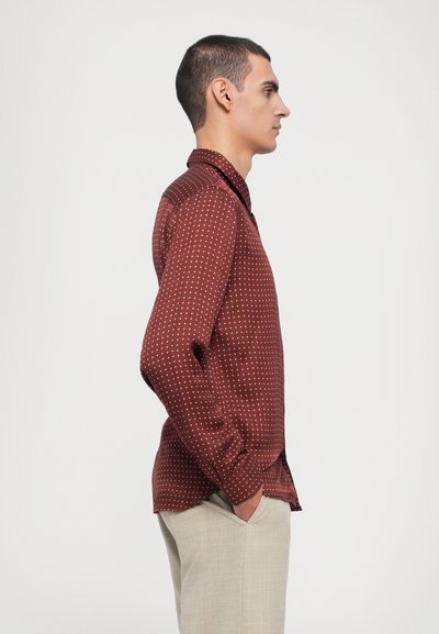 Brown button-up shirt with white polka dots, featuring a pointed collar and long sleeves, paired with beige trousers.