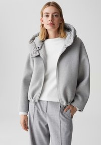 Frau trägt eine hellgraue Kapuzenjacke mit Reißverschluss, einen weißen, flauschigen Pullover und graue, maßgeschneiderte Hosen und steht vor einem schlichten Hintergrund.