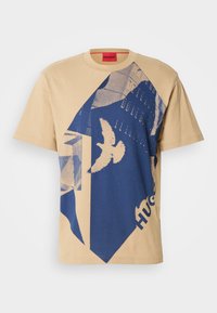 Beige bomull t-skjorte med et blått geometrisk mønster og fugledesign. Rød etikett med "HUGO" ved halsen. Korte ermer, rund hals.