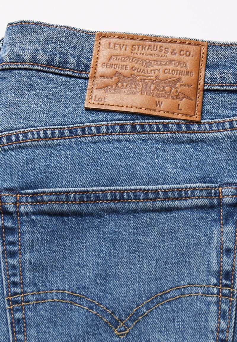 Niebieskie dżinsy z denimu z brązową skórzaną naszywką z wytłoczonym logo, widocznymi szwami i klasycznym wzorem tylnych kieszeni.