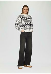 Witte trui met zwarte geometrische patronen en de tekst "MERRY", gecombineerd met zwarte wijde jeans en beige schoenen met zwarte accenten.