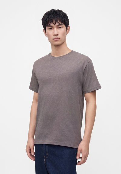 Jeune homme aux cheveux courts et foncés, portant un t-shirt gris uni à manches courtes et un jean bleu foncé sur fond blanc.