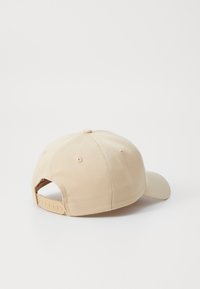Beige baseball cap med buet skjerm, stoffmateriale, justerbar bakre stropp og seks-panel design, med en sentral knapp og ventilasjonshull.