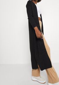 Langer schwarzer Cardigan aus Rippstrick, offenes Frontdesign, taillierte Ärmel, kombiniert mit beigen Ripp-Hosen und hellen Sneakers.