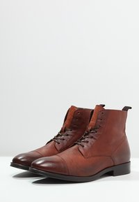 Stivali ankle in pelle marrone con finitura testurizzata, punta arrotondata e chiusura con lacci. Presentano un tacco leggero e pannelli superiori marroni a contrasto.