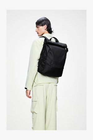 Fekete, görgős tetejű hátizsák texturált vászonból, állítható pántokkal, elülső cipzáras zsebbel és oldalsó hálós részletekkel, világos zöld ruházat fölött viselve.