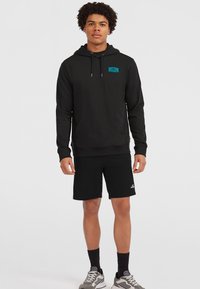 Schwarzer Hoodie mit einer Fronttasche und verstellbarem Kordelzug. Kombiniert mit schwarzen Shorts. Verfügt über ein blaues Logo auf der Brust. Graue Sneakers.