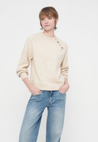 Bež pulover pleten z okroglo ovratnico, z rebrastimi manšetami in štirimi okrasnimi gumbi na levi rami. Kombiniran s svetlo modrimi širokimi hlačami.