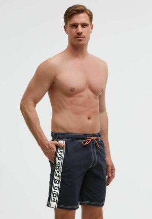 Shirtless man wearing navy blue swim shorts with a white side stripe and orange drawstring, standing with one hand in pocket.