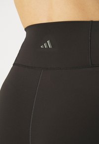 Czarne legginsy z wysokim stanem o gładkiej fakturze, z małym, wypukłym ciemnoszarym logo w postaci trzech pasków na pasie.