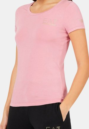 Donna che indossa una maglietta aderente rosa chiaro a maniche corte con logo di strass sul petto e pantaloni neri, una mano in tasca.