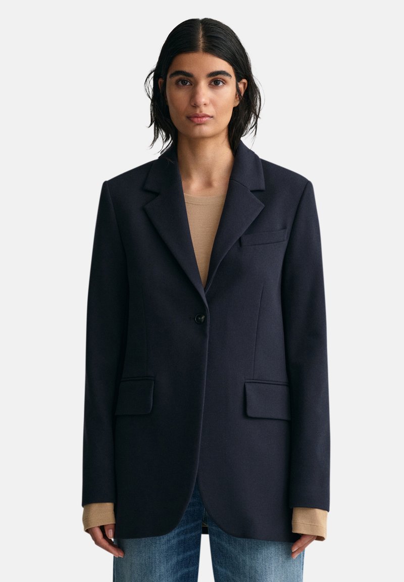 Eine Frau mit dunklen Haaren trägt ein navyblaues Blazer über einem beigen Langarmshirt und blauen Jeans und steht vor einem schlichten hellen Hintergrund.