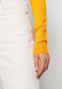 Żółta prążkowana bluzka z długim rękawem o dopasowanym kroju, w zestawieniu z białą dżinsową spódnicą. Ręka delikatnie opiera się na szwie bocznym spódnicy.