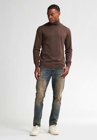Brun polotröja, figursydd design, mjuk textur; kombinerad med blekta blå jeans och vita sneakers, stående mot en ljus bakgrund.