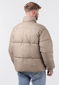 Beige Daunenjacke mit hohem Kragen, gestepptem Design und gerippten Bündchen. Weiche, glatte Textur; rechteckige Form mit horizontaler Naht.