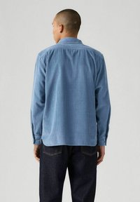 Camicia di velluto a coste blu con maniche lunghe, polsini abbottonati e colletto. Presenta una texture a coste verticale e un orlo dritto. Indossata con jeans scuri.