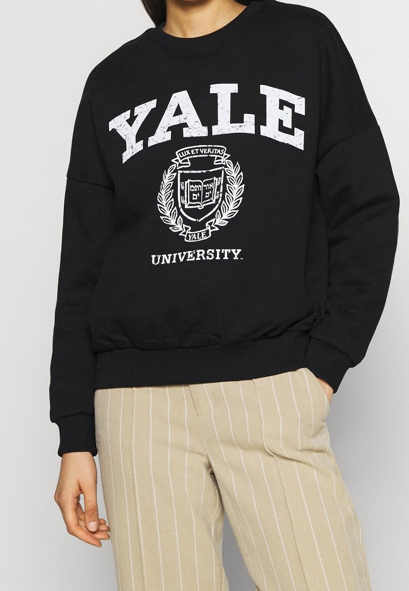 Felpa nera con la scritta "YALE" in grandi lettere bianche, arricchita con lo stemma universitario e grafiche di alloro. Maniche e orlo a costine.
