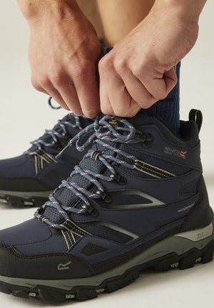 Handen die veters strikken aan marineblauwe waterdichte wandelschoenen met robuuste zolen en enkelondersteuning, gedragen met donkere sokken op een lichte achtergrond.