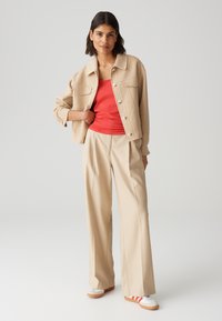 Giacca beige corta con due tasche sul petto, abbinata a una canotta rossa e pantaloni beige a gamba larga. Scarpe bianche con strisce arancioni.