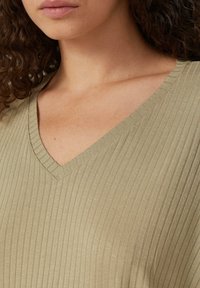 V-hals, geribbelde top in licht olijfgroene stof. Bevat een textuurpatroon en zachte drape. Toont detail van de bovenborst en de halslijn.