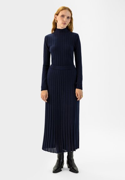 Mornarska pletena haljina s visokim ovratnikom i dugim rukavima; naborani dizajn koji seže do gležnjeva; nosi se s crnim čizmama na petu.