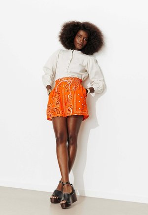 Femme avec un afro volumineux portant un chemisier blanc, un short orange vif à motif paisley et des sandales plateformes noires, appuyée contre un mur blanc.