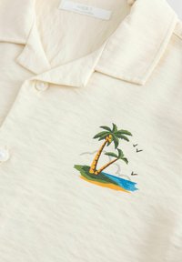 Polo shirt crème clair en tissu texturé, décoré d'un design brodé d'un palmier et d'une plage aux couleurs vives.