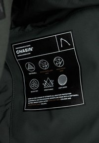 Etiqueta de tela negra unida a una chaqueta de rendimiento, con íconos y texto blancos que destacan sus propiedades de softshell, repelencia al agua y resistencia al viento.