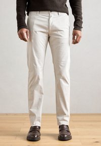 Valged puuvillased püksid sirge lõikega, millel on nööpkinnitus ja küljetaskud, kokku sobitatud pruunide nahkkingadega puidupõrandal.