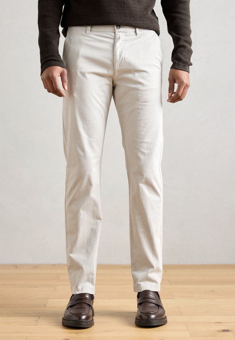 Valged puuvillased püksid sirge lõikega, millel on nööpkinnitus ja küljetaskud, kokku sobitatud pruunide nahkkingadega puidupõrandal.