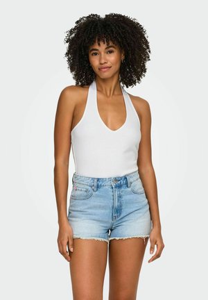 Vrouw met krullend haar die een witte haltertop en lichtblauwe spijkershorts draagt, staand tegen een effen lichte achtergrond.