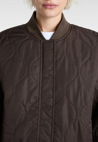 Brązowa pikowana kurtka z prążkowanym kołnierzem i pionowymi szwami. Gładka tekstura z delikatnym falistym wzorem na tkaninie.