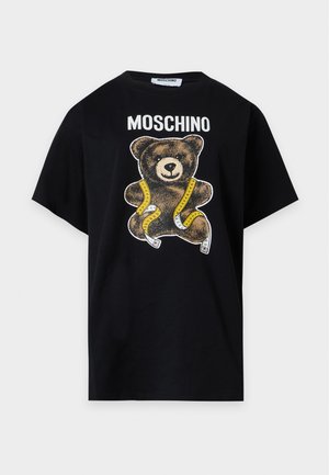 Modelka w czarnej koszulce Moschino z grafiką misia i szarych, szerokich spodniach z rękami w kieszeniach.