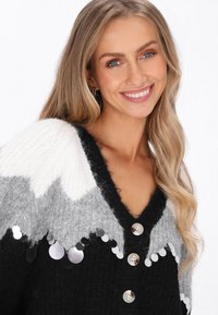 Svart, vit och grå stickad cardigan med djup V-ringning, prydd med stora knappar och en dekorativ paljettkant längs nederkanten.