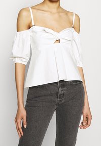 Donna che indossa una blusa bianca con spalle scoperte, maniche a palloncino e dettaglio annodato, abbinata a jeans grigi scuri a vita alta.