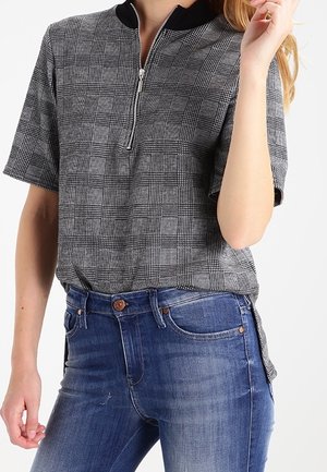 Kvinde iført en grå ternet kortærmet skjorte med sort krave og lynlås, kombineret med blå denimjeans.