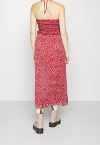 Kvinna klädd i en röd mönstrad bandeau-maxiklänning och beige snörstövlar, stående med ryggen mot kameran framför en enkel bakgrund.